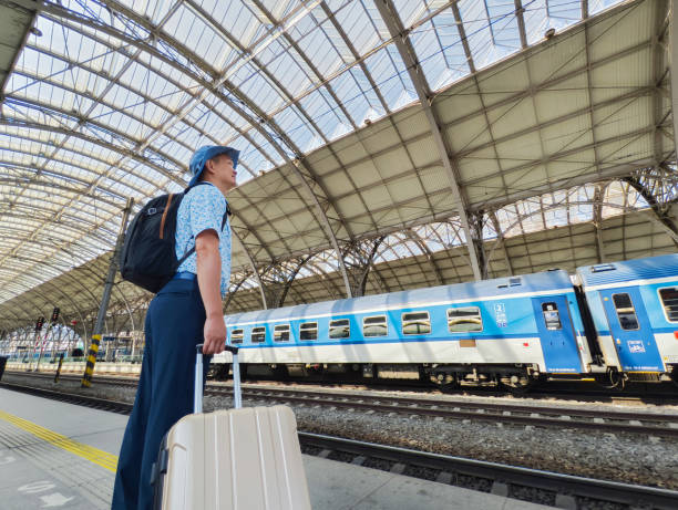 A middle-aged Asian man is waiting for the train to arrive at the train station. He is dragging his luggage, wearing a sun hat and carrying a backpack, preparing for a long journey.