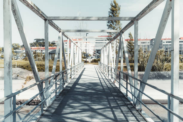 A wide-angle shot of a metal pedestrian bridge with a geometric structure of beams and railings, extending over a dry canal towards a residential area in the background, under a clear sky