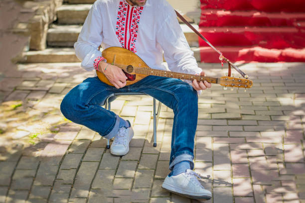 Abstract Man playing traditional folk string instrument while wearing a white embroidered shirt, seated outdoors on a sunny day