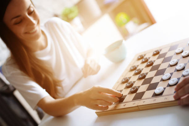 istock A smiling cute woman plays a checkers game at a white table while holding a teal mug. The scene is set in a cozy, sunlit room, conveying relaxation, joy, and a casual, playful atmosphere. 2175724465