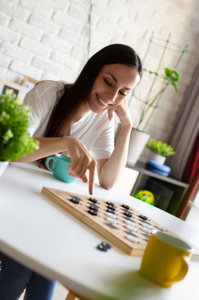 istock A young woman smiles while playing checkers at a white table. She enjoys a casual, relaxed moment in a cozy living room with a teal mug beside her. Perfect for leisure and home lifestyle themes 2175724539