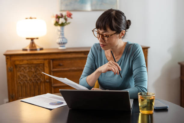 Mature Japanese woman working  from home. She is examining documents and using laptop for freelance work.