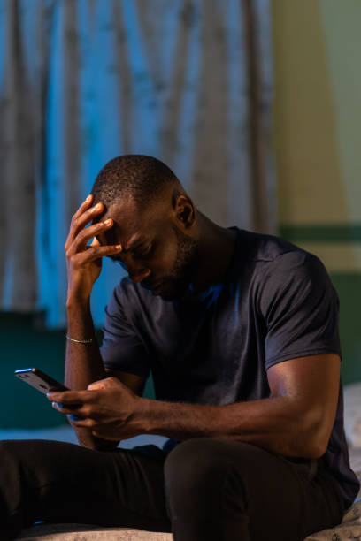 istock A young African man sits on a bed, holding his phone and looking down with a thoughtful expression. The dim lighting creates a sense of introspection and contemplation. 2175733225