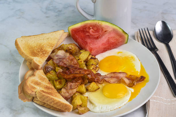 A delicious breakfast featuring two fried eggs with sunny-side up yolks, crispy bacon strips, grilled tomato, and toasted bread arranged neatly on a white plate.