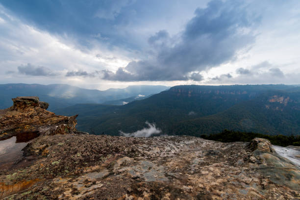 Elevated view of deep rugged valley and rolling hills from a rocky cliff edge. Gloomy day with dramatic cloudscape. White mist rising from the valley trough.