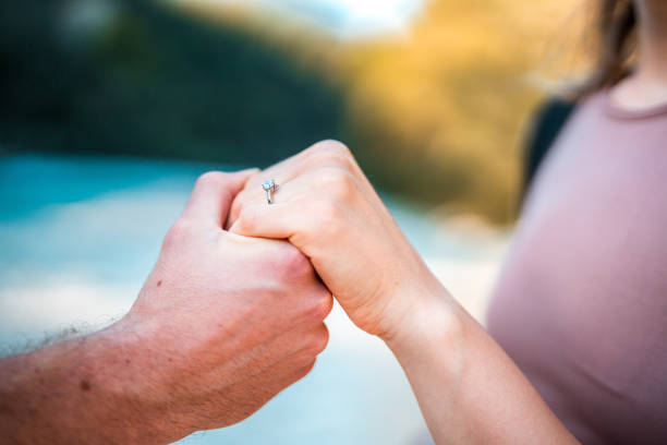 Close up on a Caucasian couple holding hands by the river, showcasing the engagement ring on the woman's hand by the river.