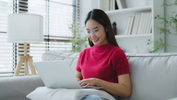 istock Happy young Asian woman using laptop while seated on couch at home 2175778546