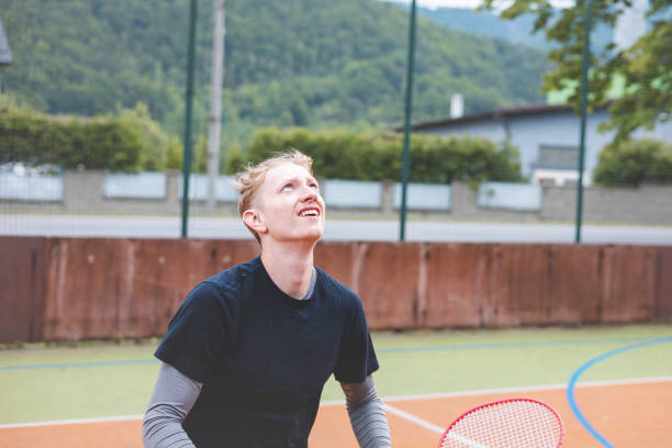 Close-up of a badminton player looking up in concentration, tracking the shuttlecock during a game. His expression shows focus and determination, framed by the natural and urban surroundings