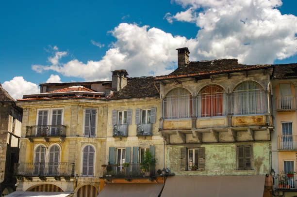 Weathered, colorful facades of traditional Italian buildings in Domodossola's main square, featuring arched windows, balconies, and shutters under a blue sky with fluffy clouds