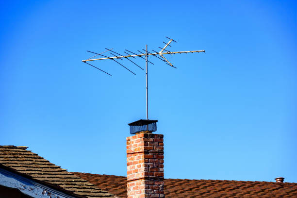 A traditional rooftop TV antenna mounted on a brick chimney rises above the brown shingle roof of a house