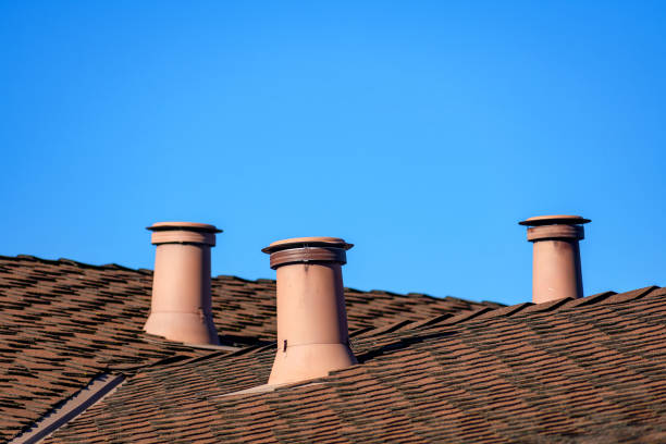 A residential roof with three vent pipes standing in a row, set against a clear blue sky. The roof is covered in brown shingles.