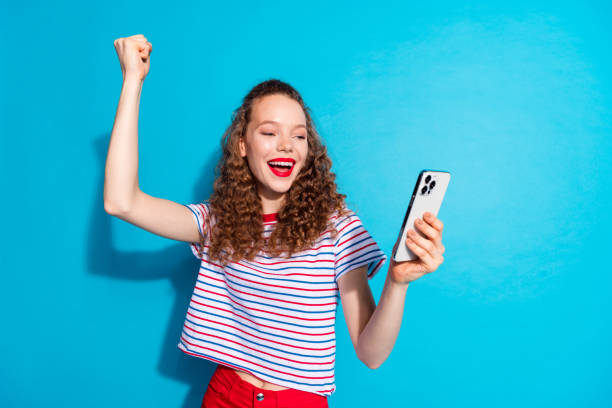istock Cheerful young woman celebrating success while looking at her smartphone with joyful expression against a vibrant blue background 2175816121