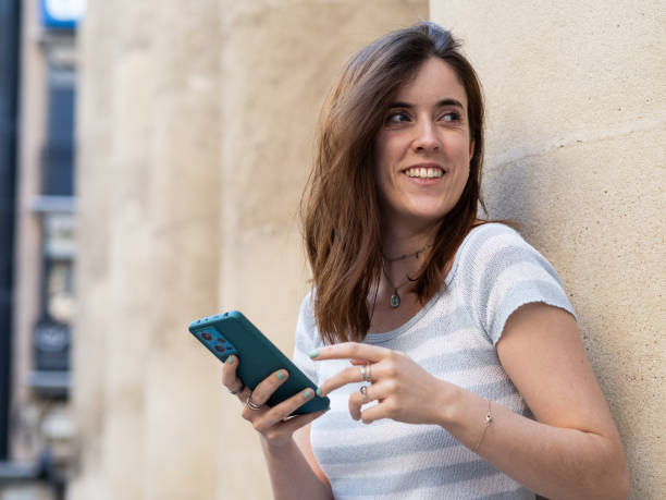istock Young woman smiling while texting on her phone against a city wall 2175817625