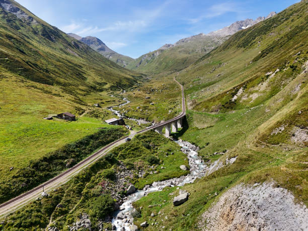 A picturesque railway bridge crossing a stream in a serene mountain valley. The lush green hills and clear blue sky provide a stunning backdrop to the peaceful landscape.
