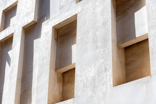 White wall with recessed windows creates an abstract pattern of light and shadow. The textured surface adds depth and dimension to the composition
