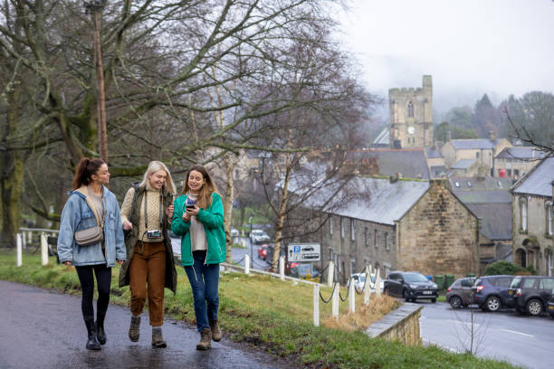 A full shot of three young female adults walking through a small village in Northumberland. They are laughing and smiling with each other, all wearing casual clothing. They are located near Rothbury, Northumberland.\n\nVideos are available similar to this scenario.