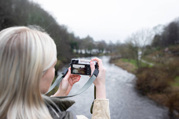 An over-the-shoulder shot of a young female adult capturing a photo of a slow-flowing river with a digital camera. She is standing in a rural village wearing outdoor walking attire. She is located near Alnwick Northumberland.\n\nVideos are available similar to this scenario.