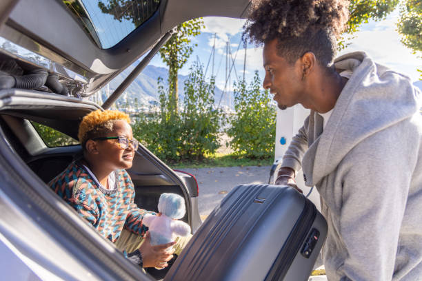 A father and son pack for a road trip. The son sits in the car trunk holding a plush toy, while the father loads luggage. A heartwarming travel moment outdoors.