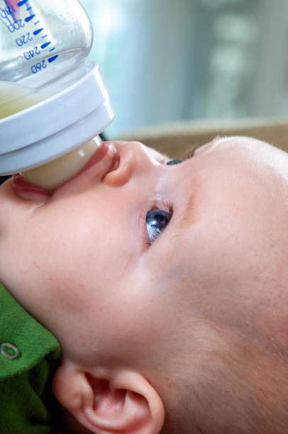 Baby drinking milk in white crib.Infant boy with formula bottle in bed. Kids nutrition.Healthy food and drink for children.Child in white nursery.Baby room interior.Kid with toy and milk bottle.Pretty baby boy drinks water from bottle lying on bed.