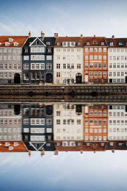 A picturesque row of colorful houses in Copenhagen lines the waterfront, their vibrant facades reflected perfectly in the calm river below. The scene is enhanced by a small boat or two gently floating in the river, adding a peaceful charm to the reflection and the classic beauty of the city’s architecture.