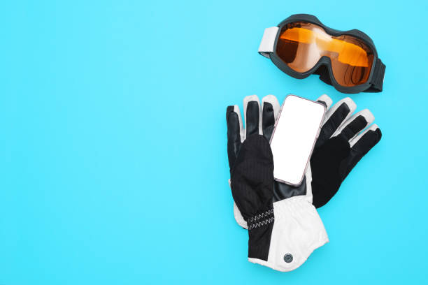 Smartphone With Blank Screen Lying On Ski Gloves With Mask On Blue Background. Flat Lay.
