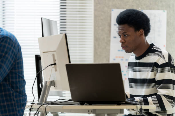 istock African-American male developers are immersed in coding and network security at a modern office. Developer is working on a laptop,  analyzes a dashboard interface. Problem-solving in a hybrid work environment, with a focus on artificial intelligence. 2175900554
