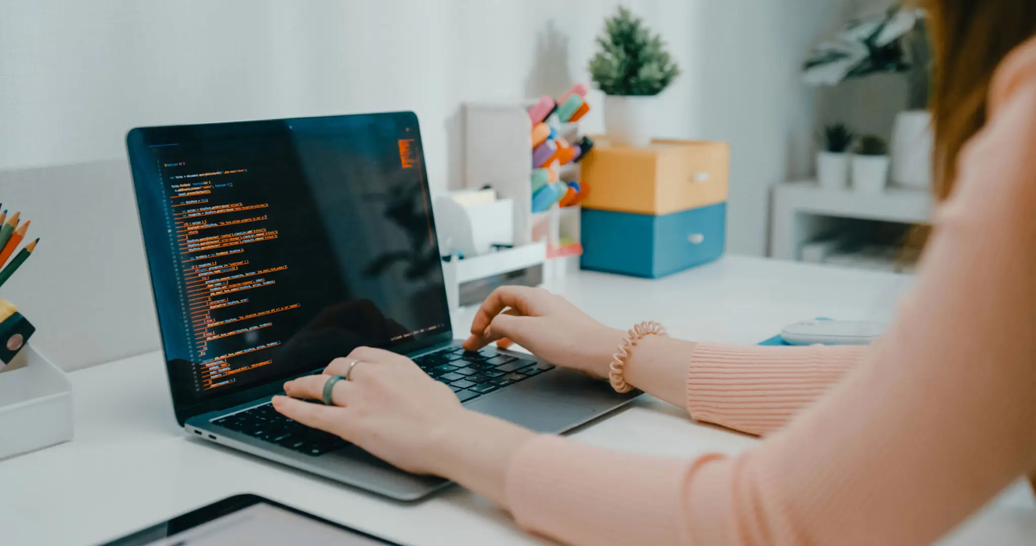 Teenager girl types code on laptop while studying at a home desk, surrounded by plants and stationery, emphasizing e-learning and technology.