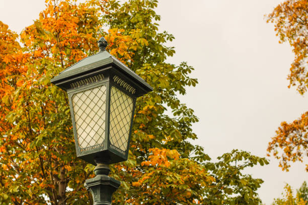 A tall lamp post stands majestically in a park, surrounded by lush green trees in the background, creating a peaceful atmosphere