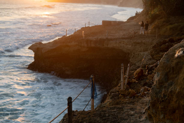 depicts a breathtaking coastal scene at sunset. The sun is setting over the ocean, casting a golden glow on the sky and water. In the foreground, there are rocky cliffs with a small platform overlooking the crashing waves. A couple is standing on the platform, enjoying the view. The overall atmosphere is peaceful and serene.