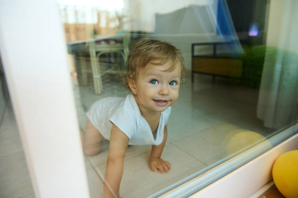 A cute baby crawls on the floor inside a home, smiling curiously through a glass door, exuding warmth and innocence.