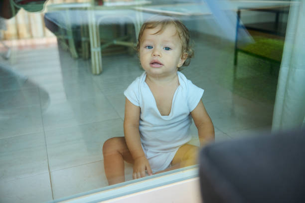 A curious toddler in a white onesie sitting on the floor, looking through a glass window. Captures curiosity, innocence, and childhood wonder in a home setting.