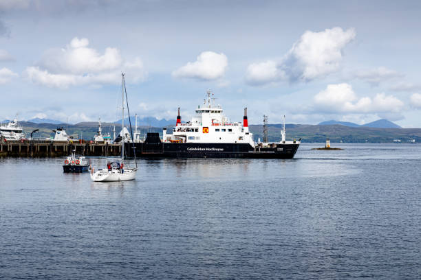 image portrays a tranquil marine environment. Dominating the scene is a large, black and white ferry, the Coruisk, aka Coir'Uisg', Caledonian MacBrayne ferry. It's docked at a pier amidst serene waters. There are several smaller boats visible, suggesting a bustling harbor area. The sky above is lightly adorned with clouds, indicating fair weather conditions. The overall scene exudes a sense of calm and order,  framed by cloudy skies and distant mountains. Ferry port of Mallaig, Lochaber, Inverness: 2024-022-07