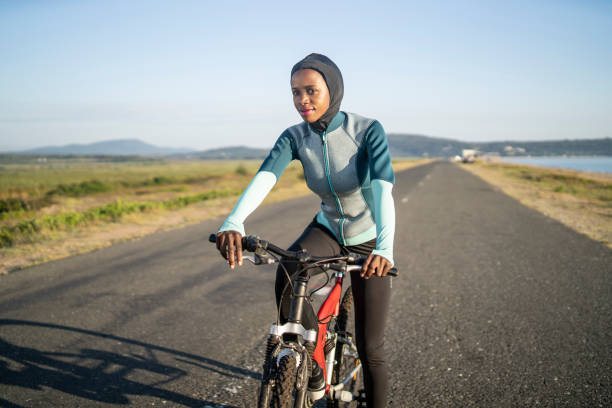 An African-American woman is riding a bike along an empty straight highway next to a narrow beach with a clear blue sky, wearing trendy cycling clothing and a hijab