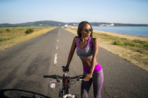 A sporty looking young woman leaning on her bike and standing in the middle of the highway on a hot summers day, wearing modern cycling clothing and sunglasses under a clear blue sky