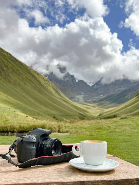 A camera rests on a table alongside a cup of coffee, with a stunning mountainous landscape in the background under a cloudy sky