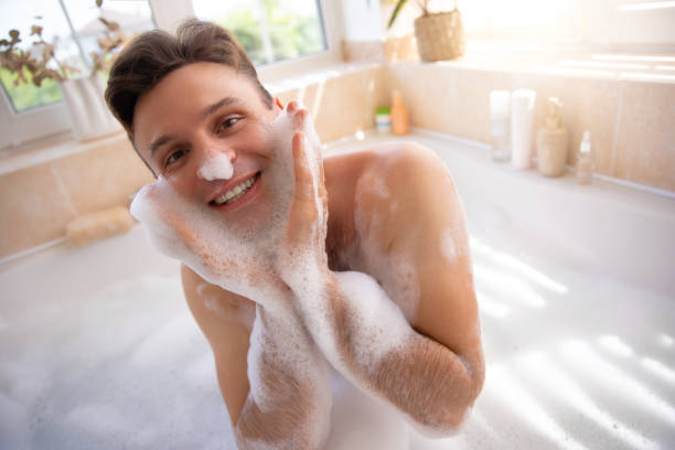 istock A joyful man playfully covers his face with bubbles in a bright, sunny bathroom. The scene, filled with natural light and plants, conveys a carefree, fun, and relaxing bath-time experience. 2175957278