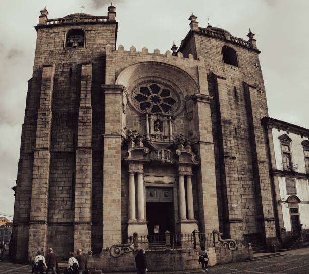 A historic cathedral with intricate architecture and people walking in front, under a cloudy sky in an urban setting.