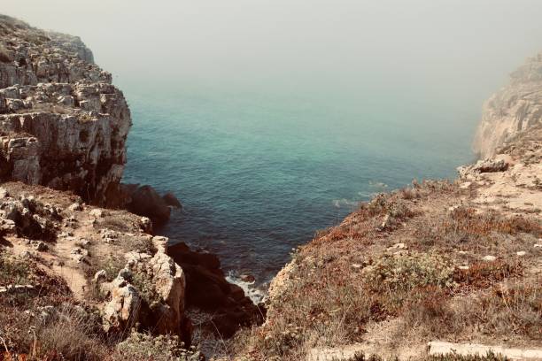 A scenic view of rocky cliffs overlooking a calm, blue ocean with a hazy sky.