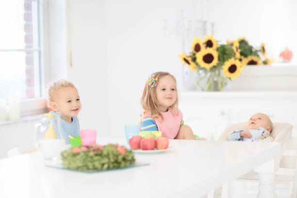 Kids eating breakfast at home. Children eat lunch in white sunny kitchen. Healthy nutrition for young child. Toddler with cereal and fruit bowl. Baby drinking milk.