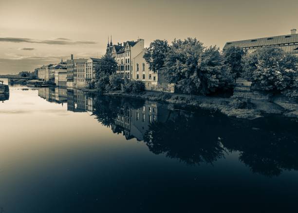 A serene riverside view with historical buildings and trees reflecting in the calm water.