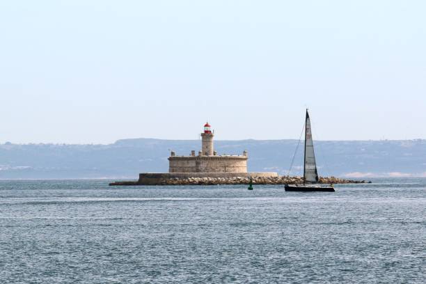 istock Sailboat passing by a historic lighthouse on a calm sea with a distant mountainous backdrop 2175974002