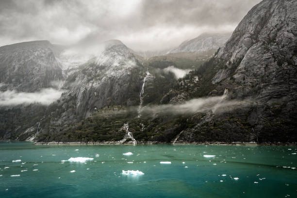 This photograph captures a rugged, mist-shrouded landscape featuring towering, rocky mountains alongside a turquoise fjord. The steep cliffs are draped with streaks of waterfalls cascading down the dark, craggy surface. Low-hanging clouds and mist wrap around the mountain slopes, contributing to the moody, dramatic atmosphere of the scene. In the foreground, floating pieces of ice are scattered across the serene, teal water, suggesting the proximity of glaciers. The combination of sharp mountain features, waterfalls, and icy water evokes a feeling of untouched wilderness, highlighting the raw and majestic beauty of nature in this remote location near Dawes Glacier, Alaska.