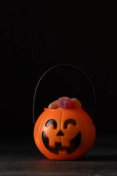 Stock photo showing close-up view of two plastic, orange, Halloween trick or treat collection buckets, shaped like pumpkin Jack O'Lanterns, filled with gummy sweets, sweet and sour jellied candy and candy corn.