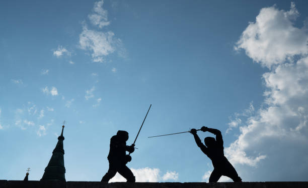 Two fencers dressed in black protective uniforms, helmets with face masks are fencing on castle wall using long medieval metal historical swords practicing before competition s. Active people concept