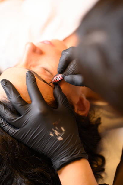Beautician applying permanent eyebrow makeup with microblading machine on brunette hispanic 30 woman