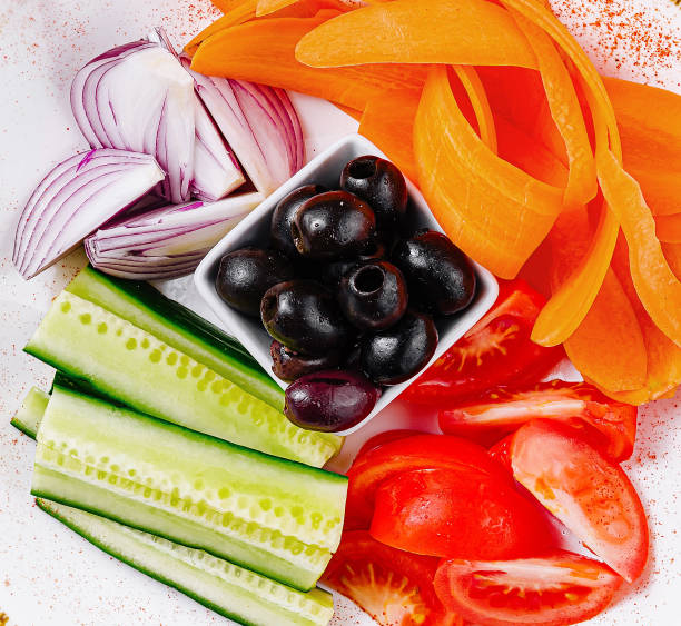 White plate presenting a colorful and healthy snack of sliced raw vegetables, including cucumber, tomatoes, red onions, and carrots, alongside a portion of black olives
