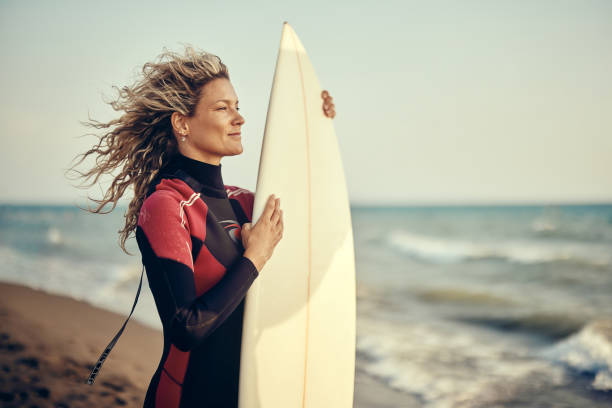Young woman with a surfboard standing on the beach and looking at waves. Copy space.