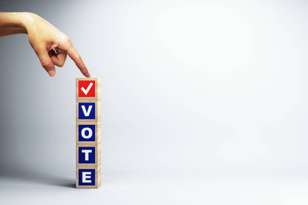 Hand pointing at wooden blocks spelling VOTE with a checkmark on a light background.