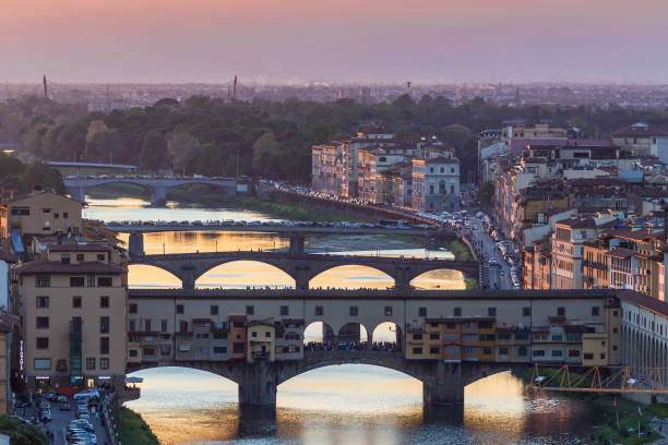 This photo, taken from Piazzale Michelangelo in Florence (Firenze), Italy, captures a stunning view of the city at sunset. The iconic Ponte Vecchio, with its historic shops lining the bridge, spans the Arno River, which reflects the warm, golden hues of the setting sun. The city's rooftops and the riverbanks create a picturesque foreground, while the distant hills and fading light provide a serene backdrop.