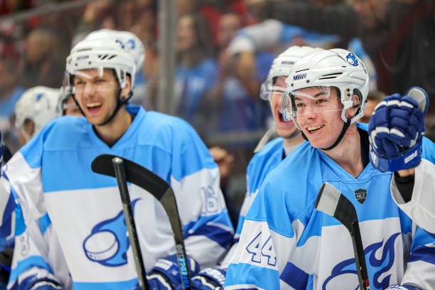 A group of hockey players wearing blue and white uniforms is captured on the bench, smiling and celebrating with enthusiasm. The players are engaged in the game, showcasing their team spirit and excitement.